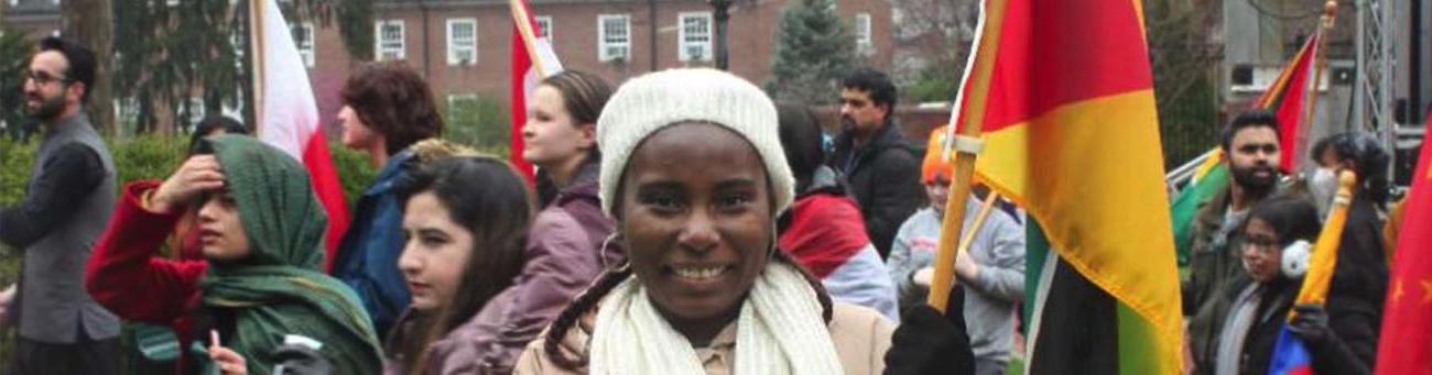 A woman wearing a white hat and scarf smiles at the camera in a crowd of people carrying international flags
