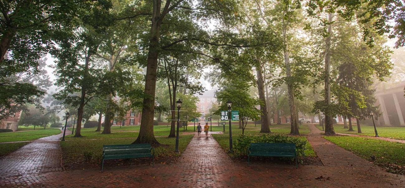 Two students are seen walking on College Green at Ohio University, surrounded by brick paths and large trees